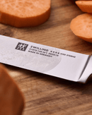 Couteau à trancher ZWILLING Four Star sur planche en bois avec légumes en arrière-plan.