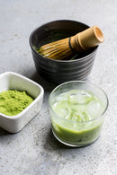 Bowl with matcha tea whisk, matcha powder, and chilled matcha drink in a glass.